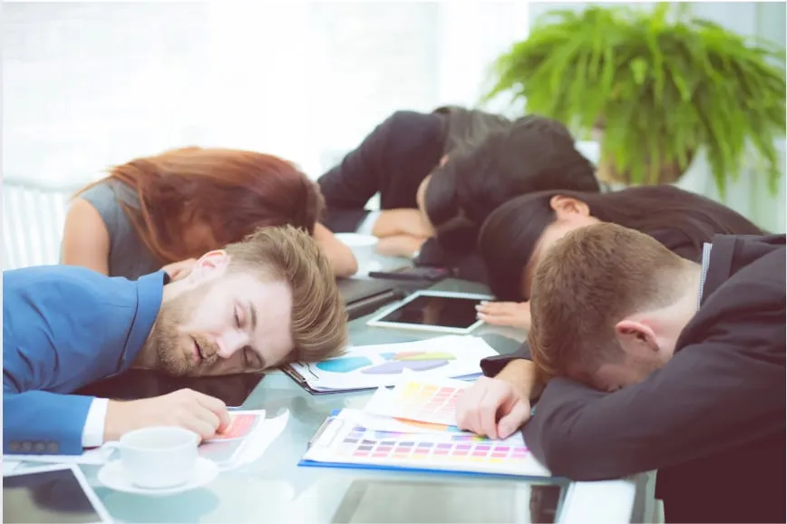 Office meeting; four people asleep at a table with documents and tablets, suggesting exhaustion. Bright room, potted plant visible.