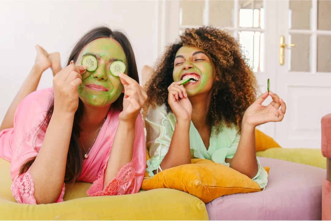 Two women laying on a bed using face masks and cucumbers on their eyes depicting "self-care"