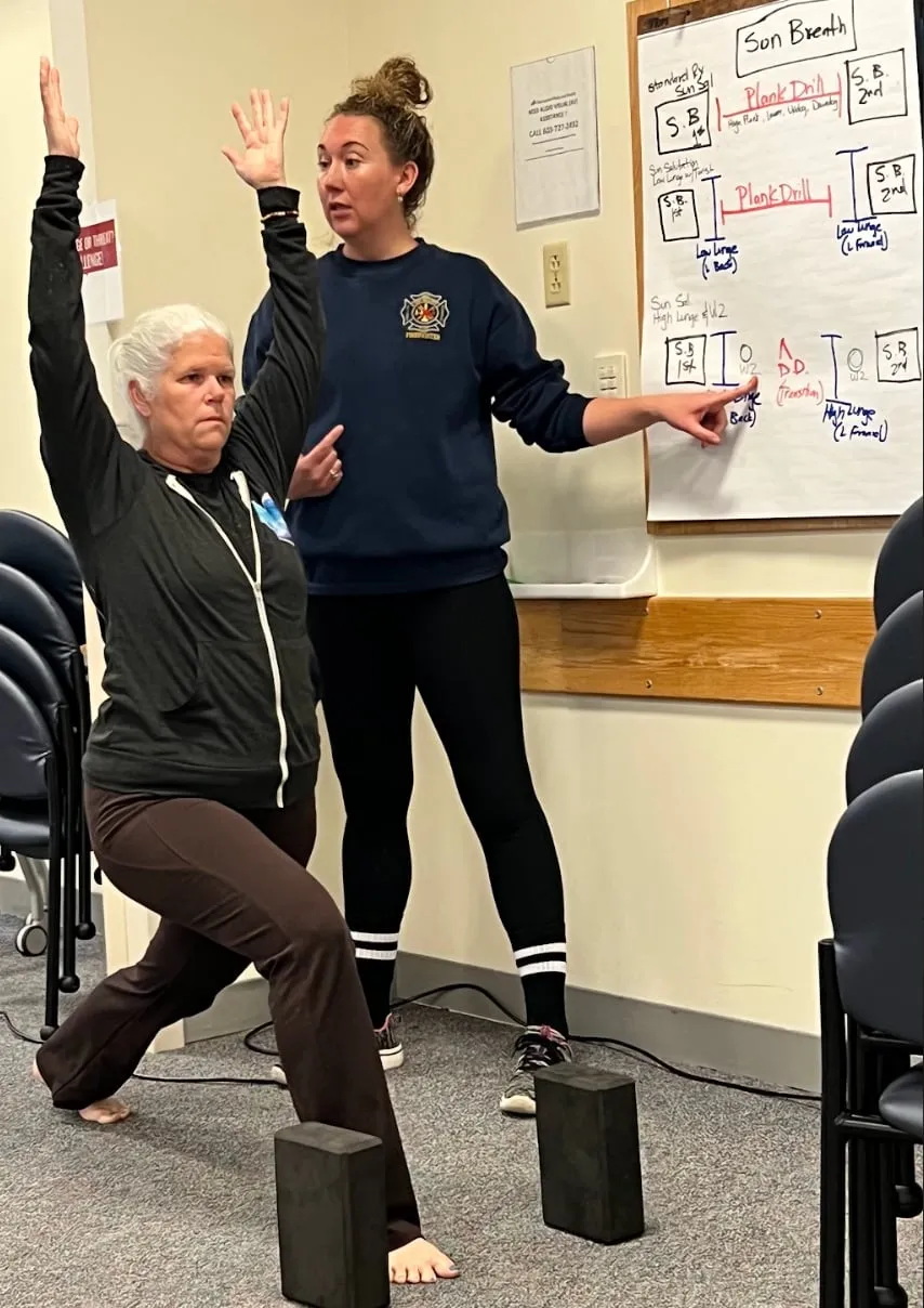 A woman in a High Lunge in front of another woman pointing to a white board with directions on it.
